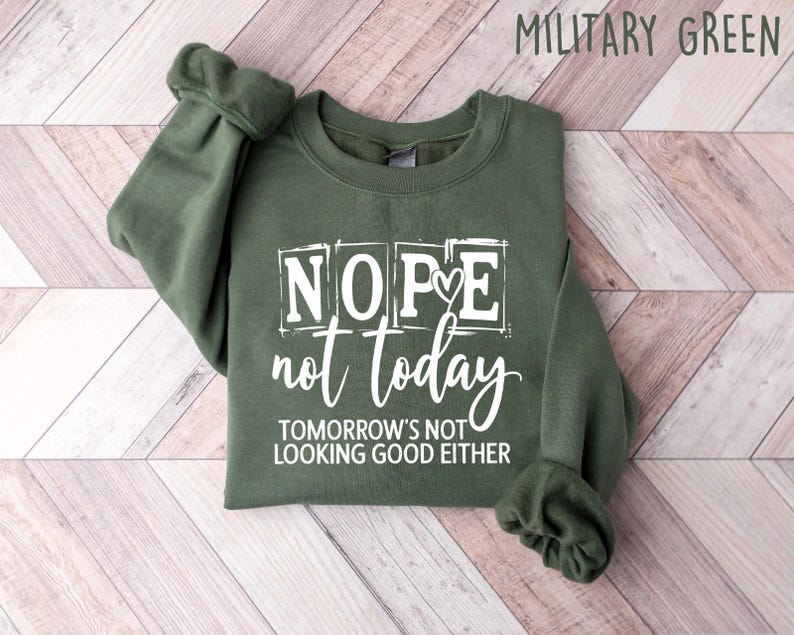 May include: Military green sweatshirt with the text "NOPE not today TOMORROW'S NOT LOOKING GOOD EITHER" in white. The sweatshirt is folded on a light wood-grain surface.