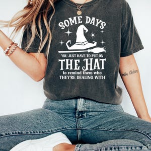 May include: Dark gray t-shirt with the text "SOME DAYS YOU JUST HAVE TO PUT ON THE HAT to remind them who THEY'RE DEALING WITH" in white. The design includes a witch hat, stars, and a broom. The shirt is worn with blue jeans and white shoes.