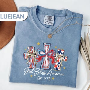 May include: Blue jean colored t-shirt with a graphic of three crosses decorated with patriotic patterns and bows. The text "God Bless America Est. 1776" is printed below the crosses. A gold necklace is draped across the shirt.