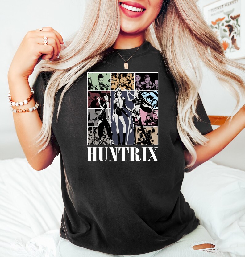 May include: Black t-shirt featuring a grid of images with a central figure in a black and white outfit. The word "HUNTRIX" is printed in white block letters below the grid. The shirt is a dark color.