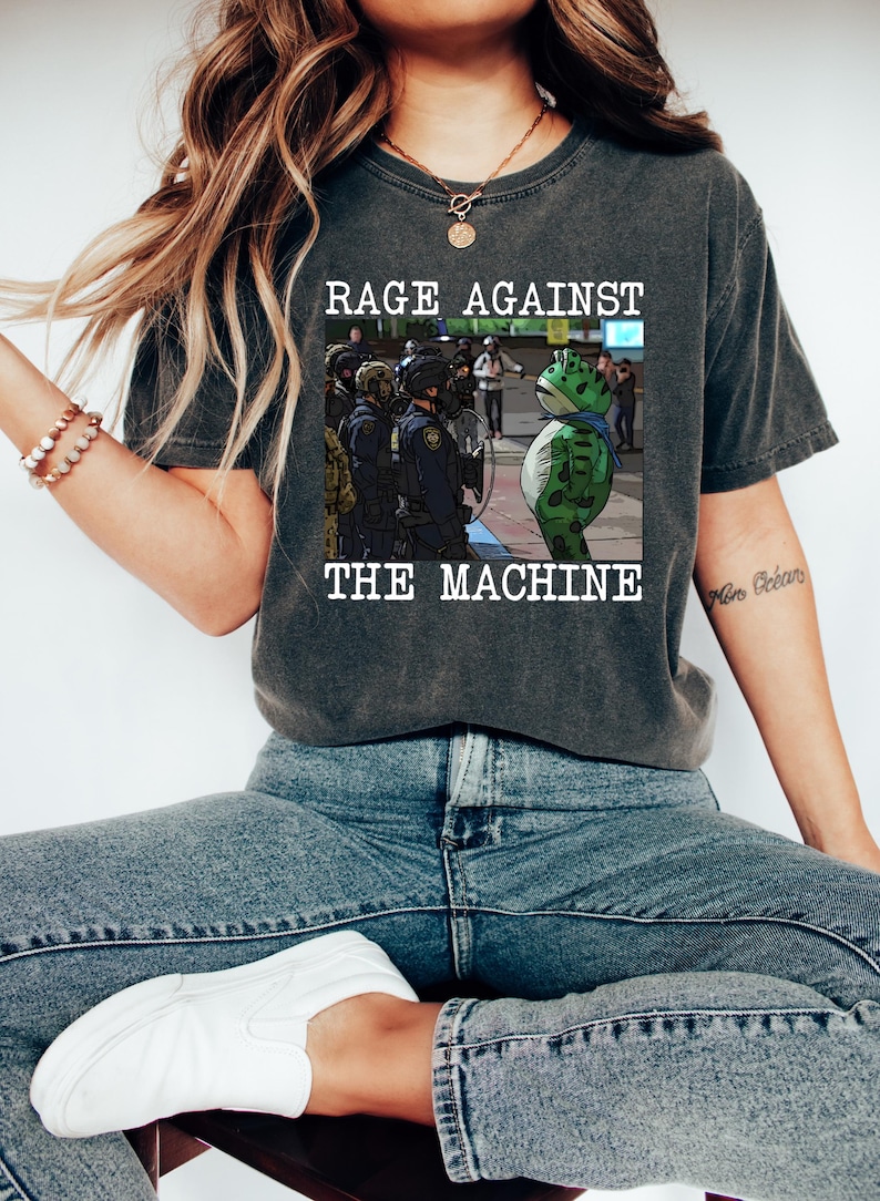 May include: Dark gray t-shirt with the text "RAGE AGAINST THE MACHINE" above an illustration of a frog facing police officers. The frog is green with a blue scarf. The shirt is worn by a person.