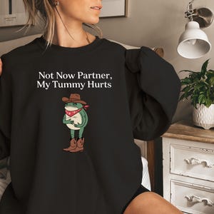 May include: Black sweatshirt with a graphic of a frog wearing a cowboy hat and bandana. The text "Not Now Partner, My Tummy Hurts" is printed on the shirt.