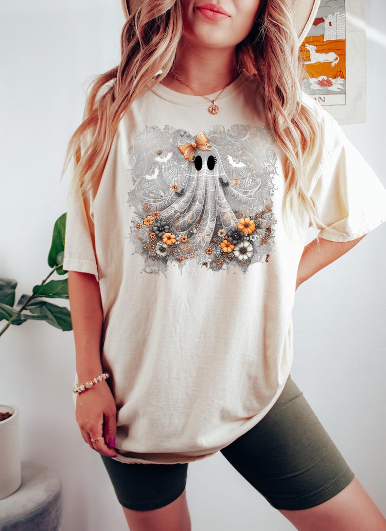 Op de afbeelding: Cr&egrave;mekleurig T-shirt met een grillig spookontwerp. Het spook heeft een strik, zwarte ogen en een vloeiend, kanten lichaam. Het ontwerp bevat bloemen en vleermuizen, perfect voor Halloween.