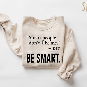 Może przedstawiać: Beżowa bluza z cytatem "Smart people don't like me." - DJT i słowami "BE SMART." pogrubioną czcionką. Rękawy są złożone. Słowo "SAND" znajduje się w prawym g&oacute;rnym rogu.