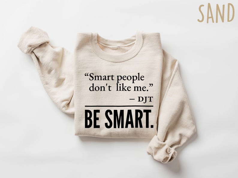 Może przedstawiać: Beżowa bluza z cytatem "Smart people don't like me." - DJT i słowami "BE SMART." pogrubioną czcionką. Rękawy są złożone. Słowo "SAND" znajduje się w prawym g&oacute;rnym rogu.