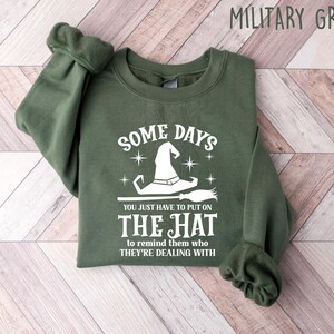 May include: Military green sweatshirt with the text "SOME DAYS YOU JUST HAVE TO PUT ON THE HAT to remind them who THEY'RE DEALING WITH". The design features a white witch hat and broom.