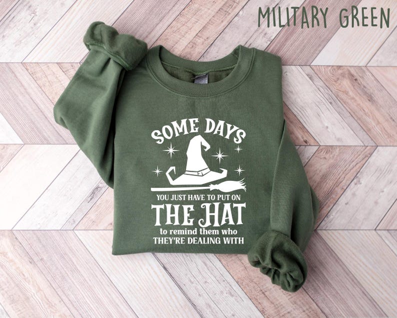 May include: Military green sweatshirt with the text "SOME DAYS YOU JUST HAVE TO PUT ON THE HAT to remind them who THEY'RE DEALING WITH". The design features a white witch hat and broom.