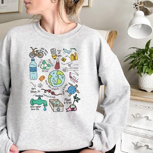 Pode incluir: Um moletom cinza claro com um gr&aacute;fico colorido com temas ambientais. O design inclui uma bicicleta, s&iacute;mbolos de reciclagem, um globo, uma tartaruga e textos como "Reciclar", "Reutilizar" e "Salvar o planeta".