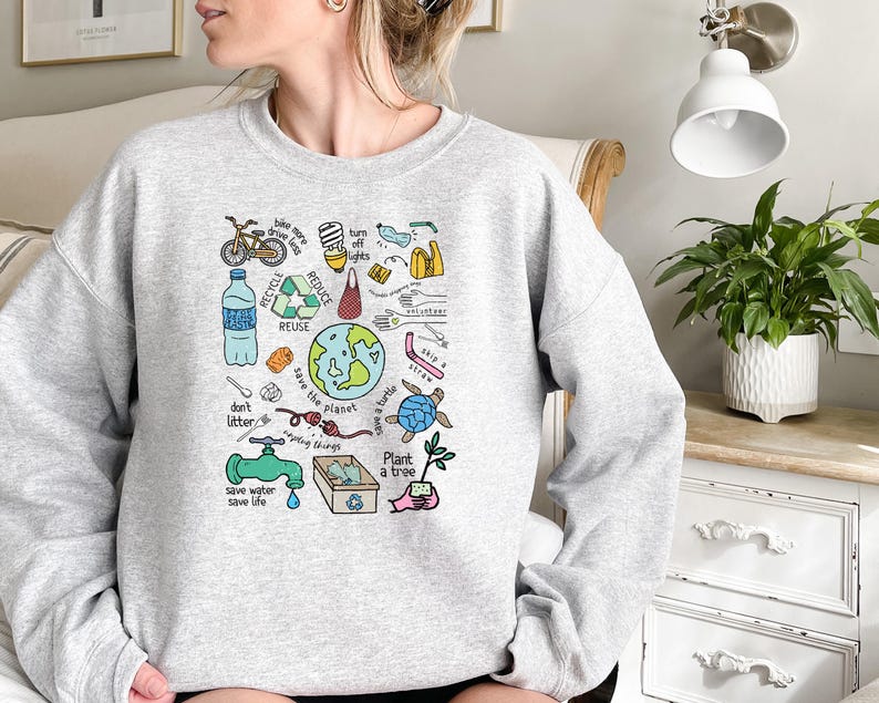 Pode incluir: Um moletom cinza claro com um gr&aacute;fico colorido com temas ambientais. O design inclui uma bicicleta, s&iacute;mbolos de reciclagem, um globo, uma tartaruga e textos como "Reciclar", "Reutilizar" e "Salvar o planeta".
