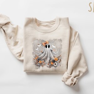 Op de afbeelding: Een zandkleurige sweatshirt met een grillig spookontwerp. Het spook heeft een strik en is omringd door bloemen en vleermuizen. De sweatshirt is gemaakt van een zacht materiaal, perfect voor Halloween of de herfst.