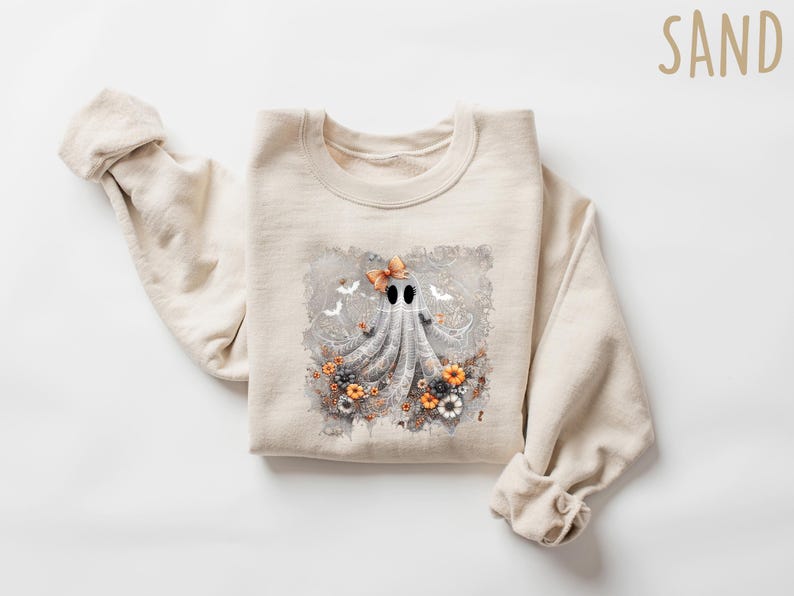 Op de afbeelding: Een zandkleurige sweatshirt met een grillig spookontwerp. Het spook heeft een strik en is omringd door bloemen en vleermuizen. De sweatshirt is gemaakt van een zacht materiaal, perfect voor Halloween of de herfst.