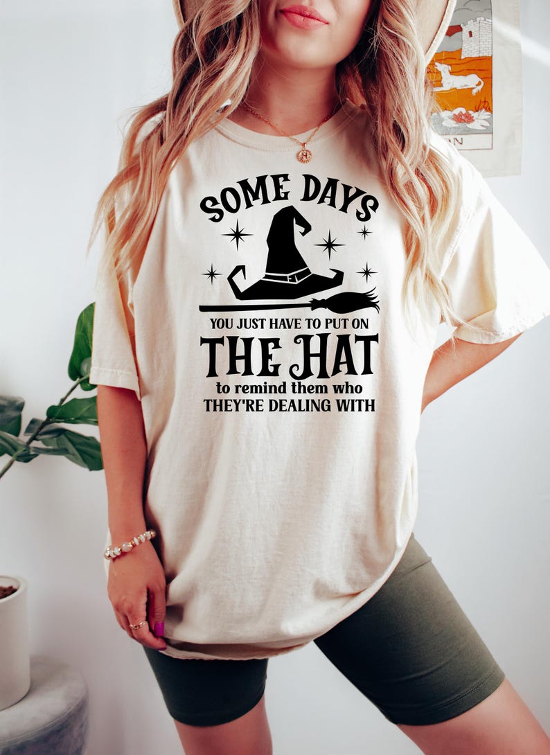 May include: Cream-colored t-shirt with a black witch hat and broom graphic. The text reads "SOME DAYS YOU JUST HAVE TO PUT ON THE HAT to remind them who THEY'RE DEALING WITH."