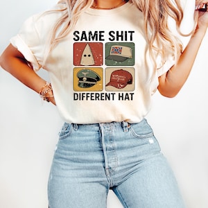 Pu&ograve; includere: T-shirt color crema con la scritta nera "SAME SHIT DIFFERENT HAT". Il design presenta quattro quadrati con immagini di diversi cappelli, tra cui un cappuccio del KKK e un cappello "Make America Great Again".