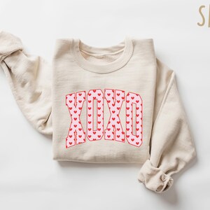 May include: A sand-colored sweatshirt with the word "XOXO" in a red and pink heart pattern. The sweatshirt is folded with the sleeves rolled up. The word "SAND" is in the upper right corner.