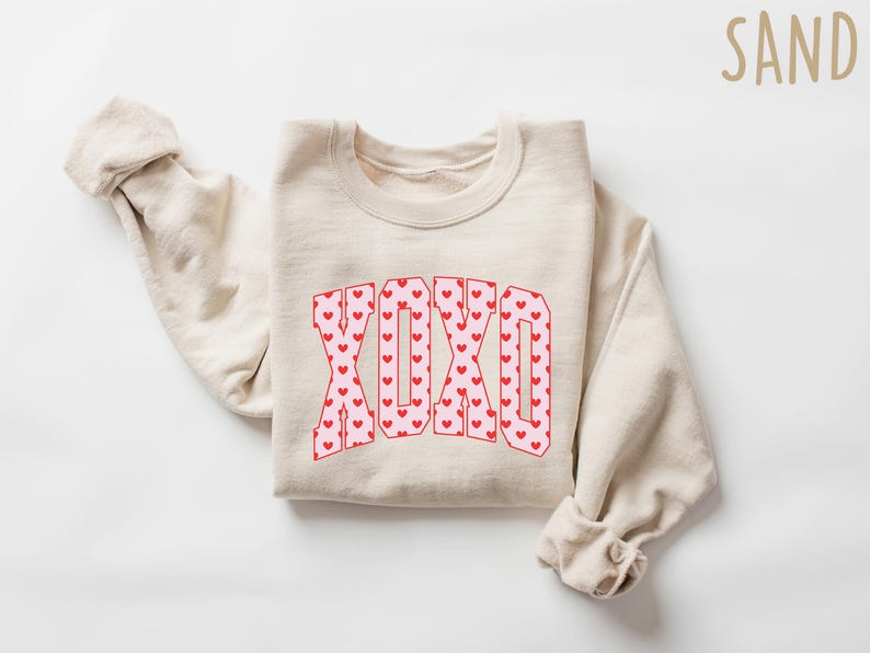 May include: A sand-colored sweatshirt with the word "XOXO" in a red and pink heart pattern. The sweatshirt is folded with the sleeves rolled up. The word "SAND" is in the upper right corner.