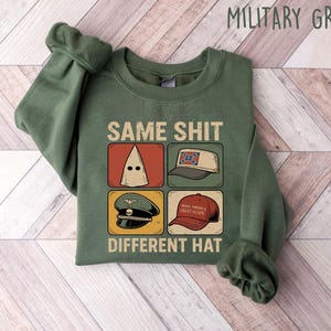 Pu&ograve; includere: Felpa verde militare con la scritta "SAME SHIT DIFFERENT HAT" e quattro illustrazioni di cappelli. I cappelli includono un cappuccio bianco, un cappello con la bandiera confederata, un cappello militare e un cappello "Make America Great Again".