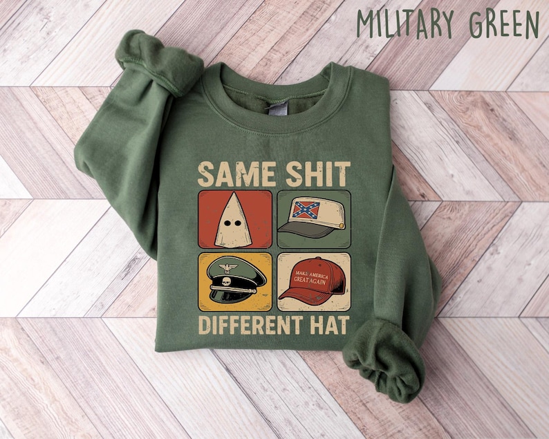 Pu&ograve; includere: Felpa verde militare con la scritta "SAME SHIT DIFFERENT HAT" e quattro illustrazioni di cappelli. I cappelli includono un cappuccio bianco, un cappello con la bandiera confederata, un cappello militare e un cappello "Make America Great Again".
