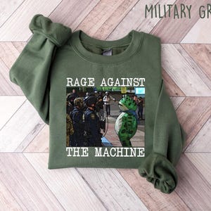 May include: Military green sweatshirt with the text "RAGE AGAINST THE MACHINE" and a graphic of a frog wearing a mask facing police officers. The sweatshirt is made of a soft material.