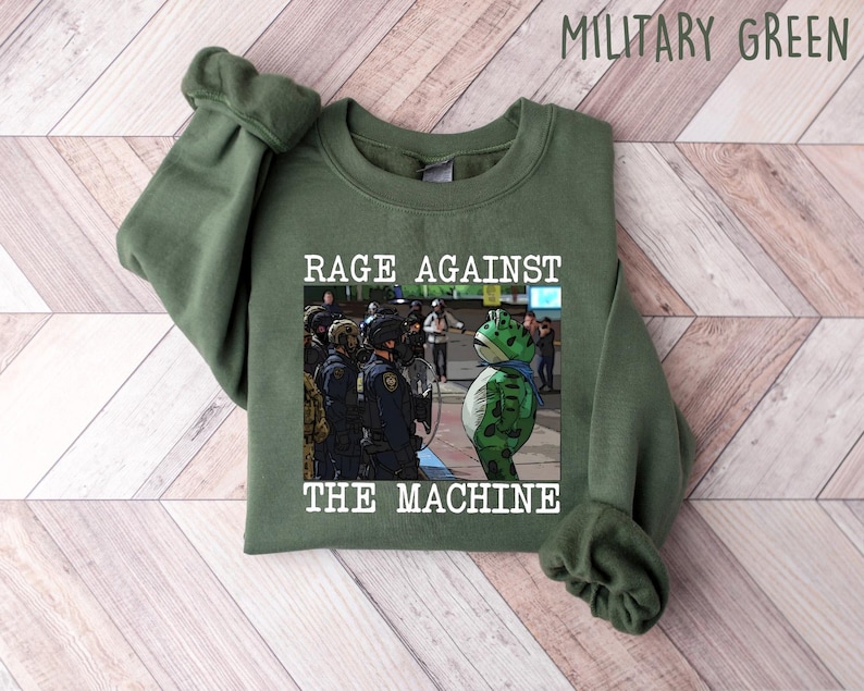 May include: Military green sweatshirt with the text "RAGE AGAINST THE MACHINE" and a graphic of a frog wearing a mask facing police officers. The sweatshirt is made of a soft material.