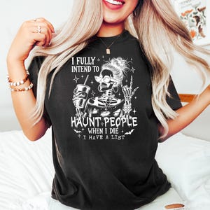 May include: Black t-shirt with a humorous Halloween graphic. The design features a skeleton with glasses, holding a drink, and making a peace sign. Text reads "I FULLY INTEND TO HAUNT PEOPLE WHEN I DIE I HAVE A LIST."