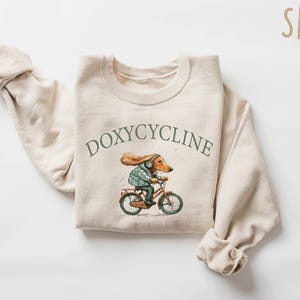 K&ouml;nnte beinhalten: Ein beiger Pullover mit einer Cartoon-Illustration eines Hundes, der Fahrrad f&auml;hrt. Der Text "DOXYCYCLINE" ist in Gr&uuml;n &uuml;ber der Illustration gedruckt. Das Wort "SAND" ist in Braun in der oberen rechten Ecke des Bildes gedruckt.