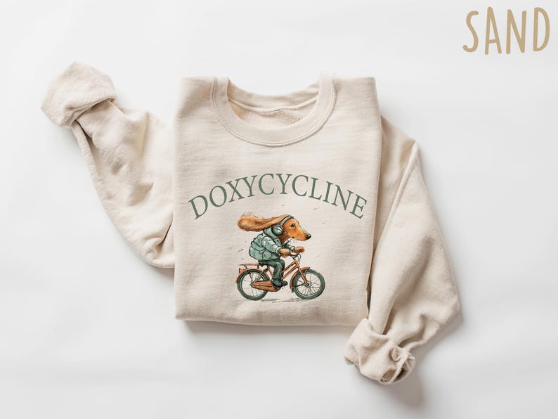 K&ouml;nnte beinhalten: Ein beiger Pullover mit einer Cartoon-Illustration eines Hundes, der Fahrrad f&auml;hrt. Der Text "DOXYCYCLINE" ist in Gr&uuml;n &uuml;ber der Illustration gedruckt. Das Wort "SAND" ist in Braun in der oberen rechten Ecke des Bildes gedruckt.