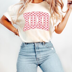 May include: Cream-colored t-shirt with the word "XOXO" in pink letters filled with red hearts. The model is wearing light blue jeans. The shirt has short sleeves and a relaxed fit.