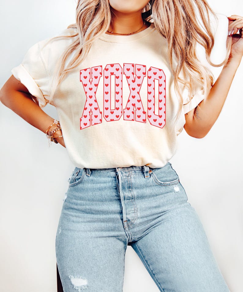 May include: Cream-colored t-shirt with the word "XOXO" in pink letters filled with red hearts. The model is wearing light blue jeans. The shirt has short sleeves and a relaxed fit.