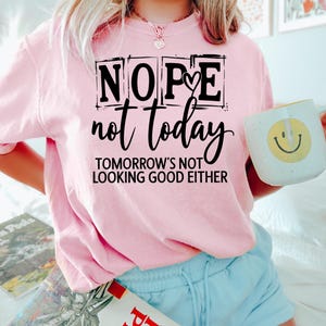 May include: A light pink sweatshirt with the words "NOPE not today TOMORROW'S NOT LOOKING GOOD EITHER" in black. A white mug with a yellow smiley face is held in the right hand. The person is wearing light blue shorts.