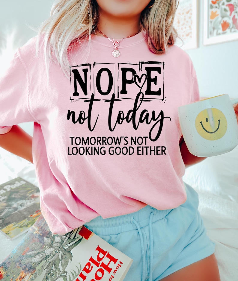 May include: A light pink sweatshirt with the words "NOPE not today TOMORROW'S NOT LOOKING GOOD EITHER" in black. A white mug with a yellow smiley face is held in the right hand. The person is wearing light blue shorts.