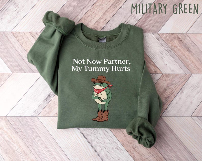 May include: A green sweatshirt with the text "Not Now Partner, My Tummy Hurts" and a cartoon frog wearing a cowboy hat and bandana.