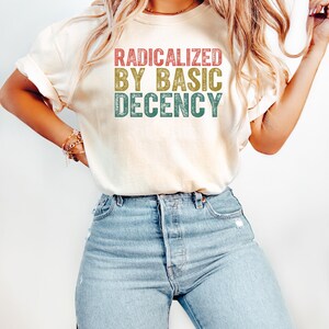 May include: A white t-shirt with the text "RADICALIZED BY BASIC DECENCY" in a vintage style font with red, yellow, and green colors.