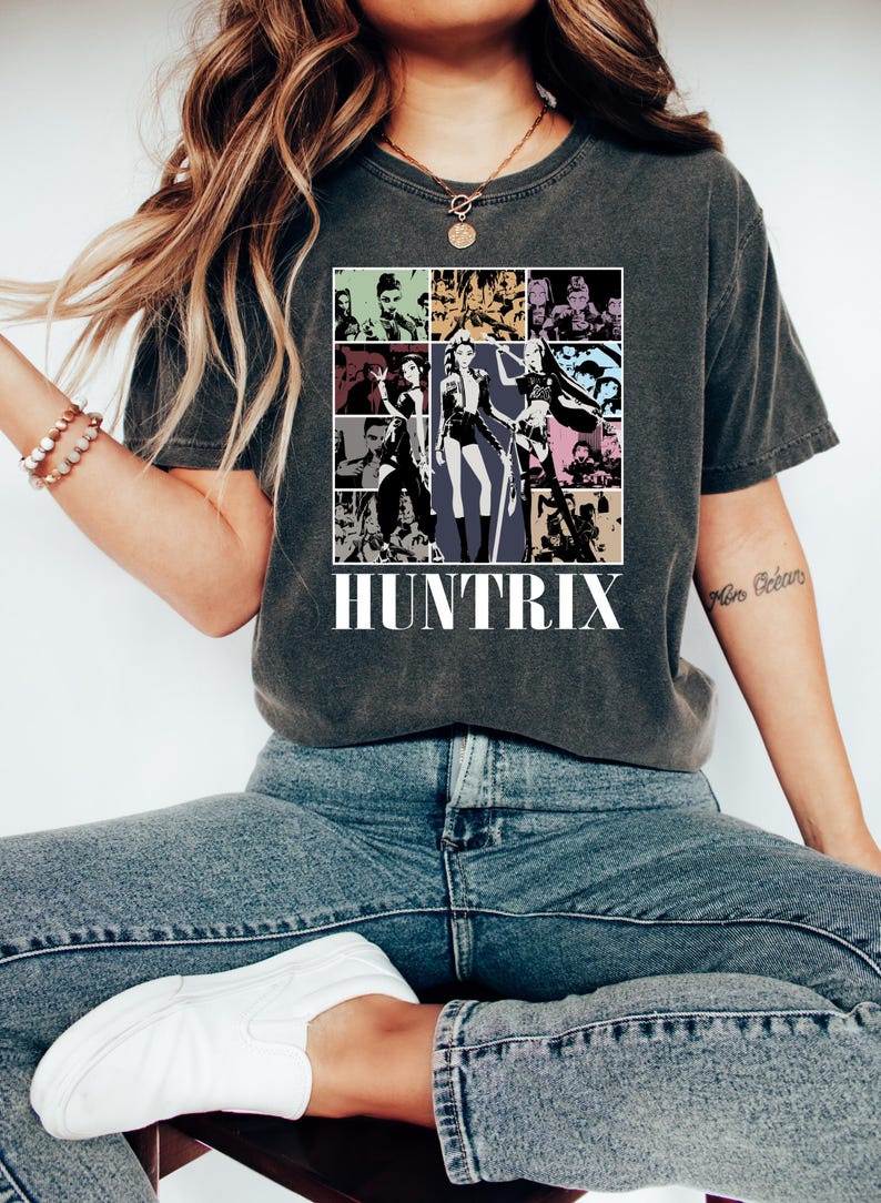 May include: Dark gray t-shirt featuring a graphic of a collage of images with the word "HUNTRIX" in white block letters. The images are in a grid pattern and feature various figures in black and white and color.