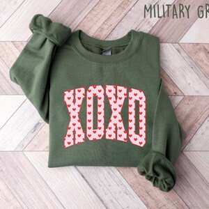 May include: A military green sweatshirt with the word "XOXO" in a pink and red heart design. The sleeves are cuffed. The text "MILITARY GREEN" is in the upper right corner. The sweatshirt is folded on a light wood surface.