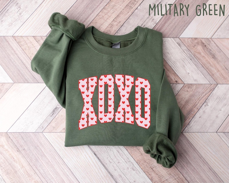 May include: A military green sweatshirt with the word "XOXO" in a pink and red heart design. The sleeves are cuffed. The text "MILITARY GREEN" is in the upper right corner. The sweatshirt is folded on a light wood surface.
