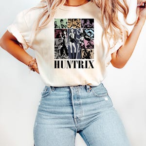 May include: Cream-colored t-shirt featuring a grid of images in various colors and styles, with the word "HUNTRIX" in bold black letters at the bottom. The shirt is paired with light blue denim jeans.