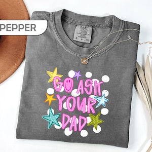 May include: A gray Comfort Colors t-shirt with the phrase "GO ASK YOUR DAD" in pink, outlined in white, and decorated with colorful stars and polka dots. A gold necklace with a pendant is on the shirt.