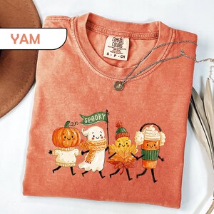 May include: A yam-colored short-sleeve t-shirt featuring a graphic of a pumpkin, a ghost holding a green flag that says SPOOKY, an autumn leaf, and a coffee cup, all with smiling faces. The shirt has a crew neckline and a Comfort Colors label.