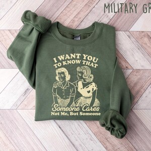 May include: A green sweatshirt with a graphic print of two women. The text on the sweatshirt reads "I want you to know that someone cares, not me, but someone".