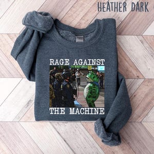 May include: Heather dark gray sweatshirt with the text "RAGE AGAINST THE MACHINE" above an image of a frog in front of police officers. The sweatshirt is a dark gray color.