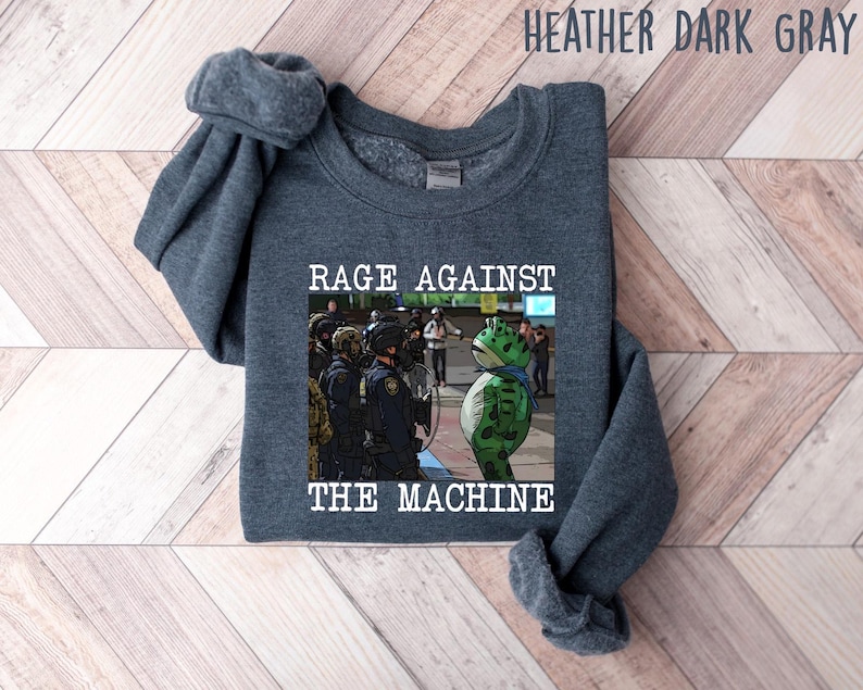 May include: Heather dark gray sweatshirt with the text "RAGE AGAINST THE MACHINE" above an image of a frog in front of police officers. The sweatshirt is a dark gray color.