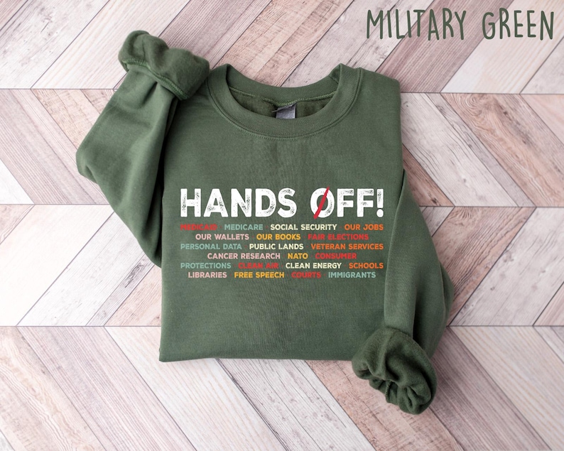Op de afbeelding: Een groen sweatshirt met de tekst "HANDS OFF!" in wit. De tekst wordt omringd door een lijst met items in rood, oranje en geel, waaronder "Medicare", "Sociale zekerheid", "Onze banen", "Onze portemonnees", "Onze boeken", "Eerlijke verkiezingen", "Persoonlijke gegevens", "Openbaar land", "Veteranendiensten", "Kankeronderzoek", "NATO", "Consumentenbescherming", "Schone energie", "Scholen", "Bibliotheken", "Vrijheid van meningsuiting", "Bezuinigingen" en "Immigranten".