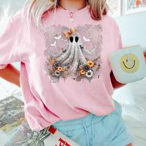 Op de afbeelding: Lichtroze T-shirt met een grillig spookontwerp. Het spook heeft een strik, omringd door bloemen en vleermuizen. Het shirt maakt deel uit van een Halloween-collectie.