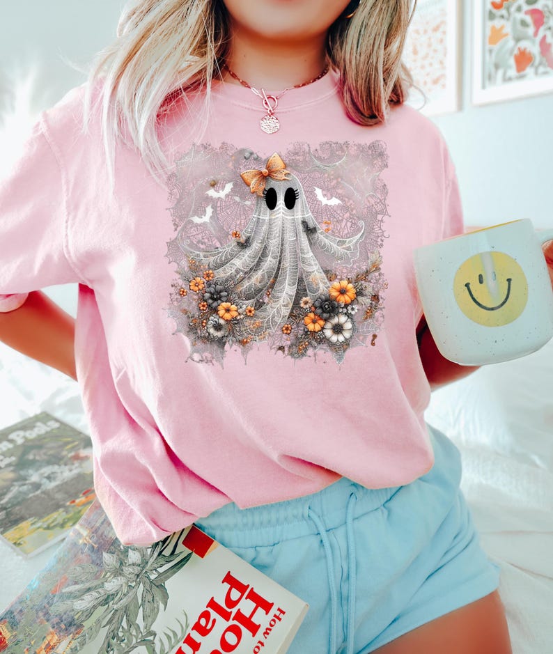 Op de afbeelding: Lichtroze T-shirt met een grillig spookontwerp. Het spook heeft een strik, omringd door bloemen en vleermuizen. Het shirt maakt deel uit van een Halloween-collectie.