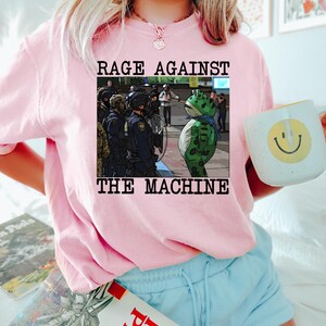 May include: A light pink sweatshirt with the text "RAGE AGAINST THE MACHINE" above an illustration of a frog in front of police officers. The person is also holding a white mug with a yellow smiley face.