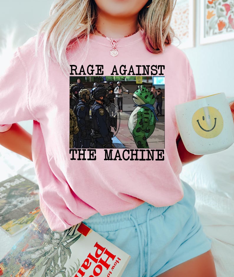 May include: A light pink sweatshirt with the text "RAGE AGAINST THE MACHINE" above an illustration of a frog in front of police officers. The person is also holding a white mug with a yellow smiley face.