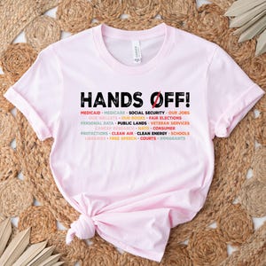 Op de afbeelding: Roze T-shirt met de tekst "HANDS OFF!" in zwart. De tekst wordt omringd door een lijst met items in verschillende kleuren, waaronder Medicaid, Medicare, Sociale Zekerheid, Onze banen, Onze portemonnees, Onze boeken, Eerlijke verkiezingen, Persoonsgegevens, Openbaar land, Veteranenvoorzieningen, Kankeronderzoek, NAVO, Consumentenbescherming, Schone lucht, Schone energie, Scholen, Bibliotheken, Vrijheid van meningsuiting, Rechtbanken en Immigranten.