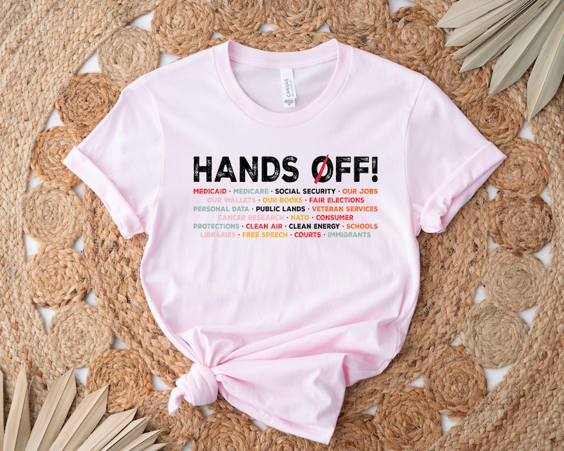 Op de afbeelding: Roze T-shirt met de tekst "HANDS OFF!" in zwart. De tekst wordt omringd door een lijst met items in verschillende kleuren, waaronder Medicaid, Medicare, Sociale Zekerheid, Onze banen, Onze portemonnees, Onze boeken, Eerlijke verkiezingen, Persoonsgegevens, Openbaar land, Veteranenvoorzieningen, Kankeronderzoek, NAVO, Consumentenbescherming, Schone lucht, Schone energie, Scholen, Bibliotheken, Vrijheid van meningsuiting, Rechtbanken en Immigranten.