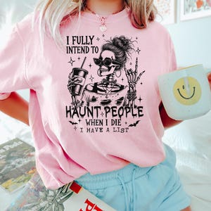 May include: A light pink t-shirt with a graphic of a skeleton wearing sunglasses, holding a drink, and making a rock-and-roll hand gesture. The text reads "I FULLY INTEND TO HAUNT PEOPLE WHEN I DIE I HAVE A LIST."