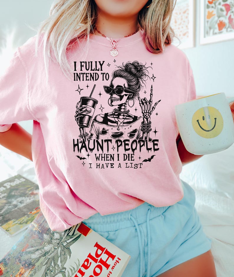 May include: A light pink t-shirt with a graphic of a skeleton wearing sunglasses, holding a drink, and making a rock-and-roll hand gesture. The text reads "I FULLY INTEND TO HAUNT PEOPLE WHEN I DIE I HAVE A LIST."